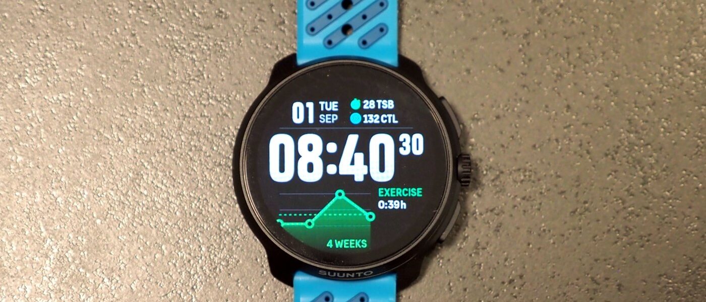 שעון Suunto Race 2 עם רצועה כחולה מונח על משטח בהיר, מסך שעון ראשי