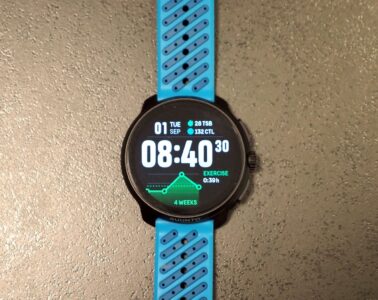 שעון Suunto Race 2 עם רצועה כחולה מונח על משטח בהיר, מסך שעון ראשי